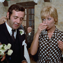 Man in suit pouring drink while woman in patterned dress sips from glass at festive indoor gathering with dancing guests.