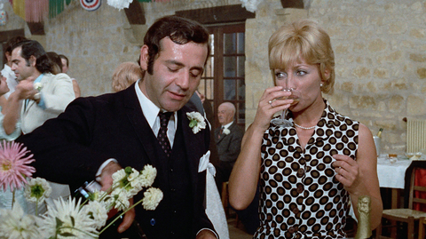 Man in suit pouring drink while woman in patterned dress sips from glass at festive indoor gathering with dancing guests.