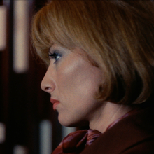 Close-up profile of a woman with short blonde hair and red lipstick, wearing a brown outfit and a patterned scarf.