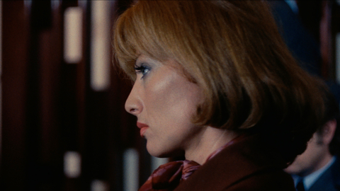 Close-up profile of a woman with short blonde hair and red lipstick, wearing a brown outfit and a patterned scarf.