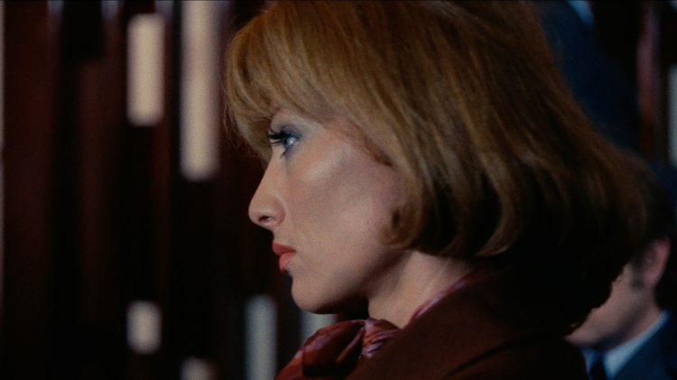 Close-up profile of a woman with short blonde hair and red lipstick, wearing a brown outfit and a patterned scarf.