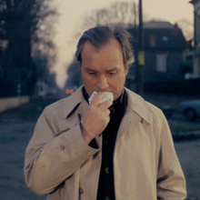 Man in a beige coat standing on a street, holding a tissue to his nose, with blurred houses and a car in the background at dusk.