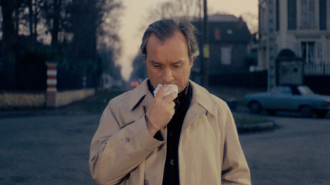 Man in a beige coat standing on a street, holding a tissue to his nose, with blurred houses and a car in the background at dusk.