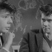 Black-and-white photo of two young men in suits, one resting chin on hand, with a chalkboard covered in drawings behind them.