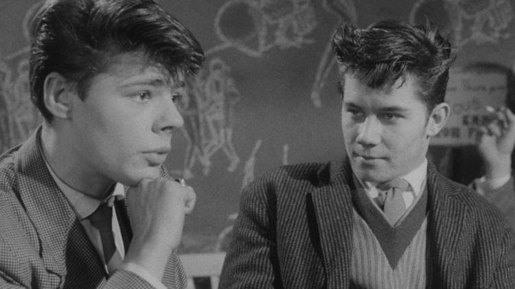 Black-and-white photo of two young men in suits, one resting chin on hand, with a chalkboard covered in drawings behind them.
