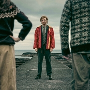Man in red jacket and tie standing on a pier facing the camera, flanked by two people wearing patterned sweaters near a cloudy sea.
