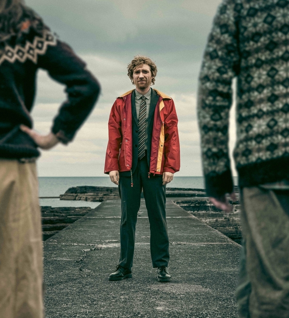 Man in red jacket and tie standing on a pier facing the camera, flanked by two people wearing patterned sweaters near a cloudy sea.