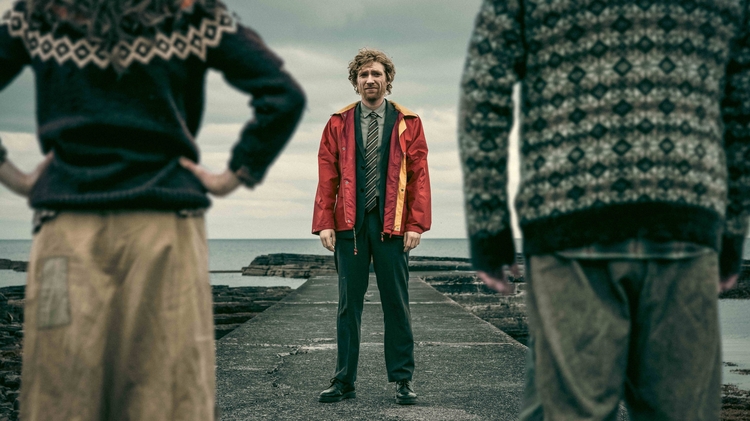 Man in red jacket and tie standing on a pier facing the camera, flanked by two people wearing patterned sweaters near a cloudy sea.