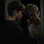 Two young men separated by a screen door, touching hands and looking solemnly at each other in dim lighting.