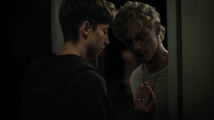 Two young men separated by a screen door, touching hands and looking solemnly at each other in dim lighting.