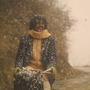 Man wearing a scarf and coat joyfully riding a bicycle on a snowy, winding mountain road with trees in the background.