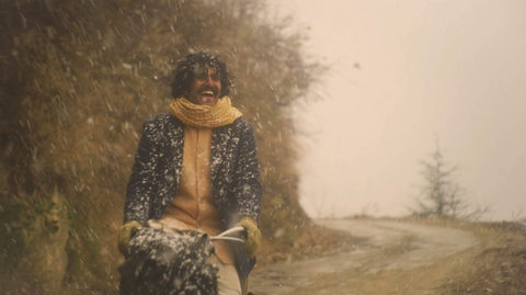 Man wearing a scarf and coat joyfully riding a bicycle on a snowy, winding mountain road with trees in the background.