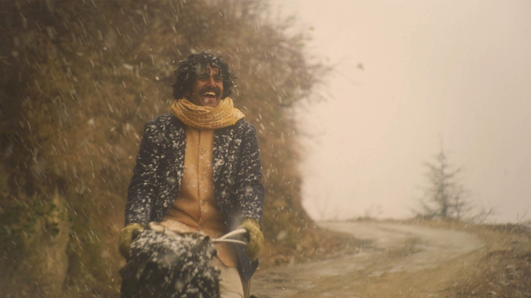 Man wearing a scarf and coat joyfully riding a bicycle on a snowy, winding mountain road with trees in the background.