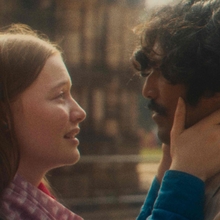 A woman tenderly holds a man's face as they gaze at each other outdoors with a blurred background.