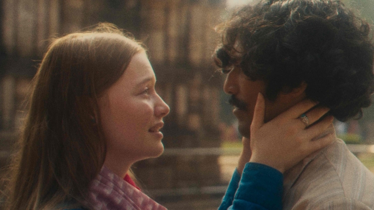 A woman tenderly holds a man's face as they gaze at each other outdoors with a blurred background.