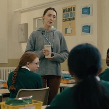 Teacher in a grey sweater holds a cup, talking to attentive students in green uniforms seated in a classroom.