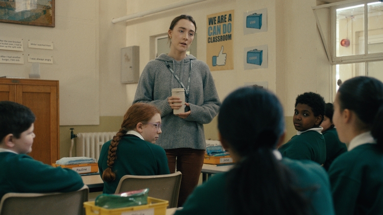 Teacher in a grey sweater holds a cup, talking to attentive students in green uniforms seated in a classroom.