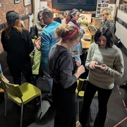 Adults gather in a cozy brick-walled workshop with string lights, chatting around tables and chairs; a screen and colorful artwork create a creative mood.