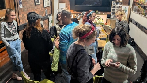 Adults gather in a cozy brick-walled workshop with string lights, chatting around tables and chairs; a screen and colorful artwork create a creative mood.