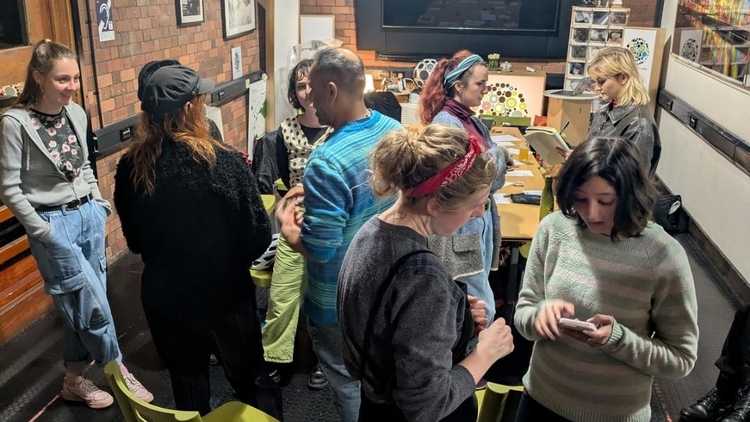 Adults gather in a cozy brick-walled workshop with string lights, chatting around tables and chairs; a screen and colorful artwork create a creative mood.