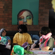 A performer in a red velvet coat and feathered hat chats with seated guests at a colorful art table beneath a bold painted portrait.