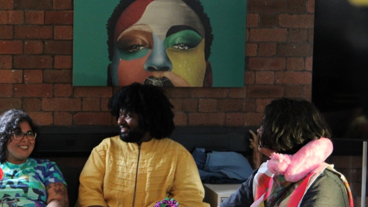 A performer in a red velvet coat and feathered hat chats with seated guests at a colorful art table beneath a bold painted portrait.