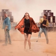 Three people stand in a dusty desert with large speakers behind them, surrounded by a cloud of dust.
