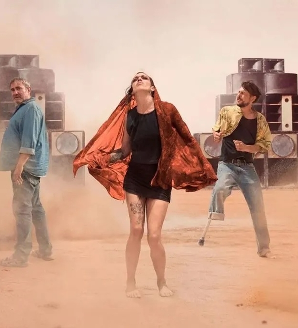 Three people stand in a dusty desert with large speakers behind them, surrounded by a cloud of dust.