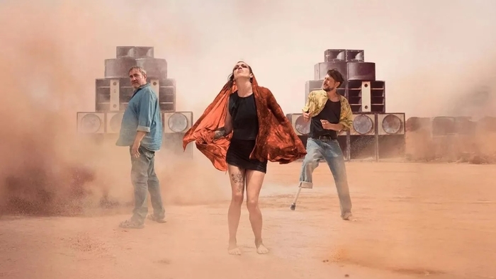 Three people stand in a dusty desert with large speakers behind them, surrounded by a cloud of dust.