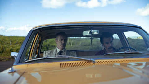 Two men sit quietly inside a yellow vintage car on a rural road under a blue sky with scattered clouds.