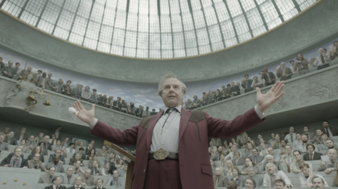 Man in maroon suit with arms raised addresses a large audience in a domed, ornate auditorium with a glass ceiling.