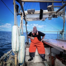 Fisherman in orange overalls sitting on a boat holding a cup, with ocean and blue sky in the background.