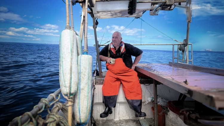 Fisherman in orange overalls sitting on a boat holding a cup, with ocean and blue sky in the background.