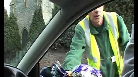 Person in a safety vest handing a loaf of bread to someone inside a car near a stone building with greenery.
