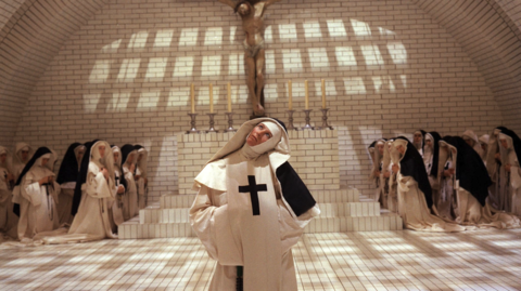 Nuns in white and black habits gathered in a tiled chapel, one nun in front with a large cross looks upward beneath a crucifix.
