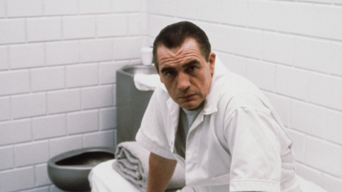 Man in white uniform sitting on a bench in a small, white-tiled room with folded towels and a metal toilet.