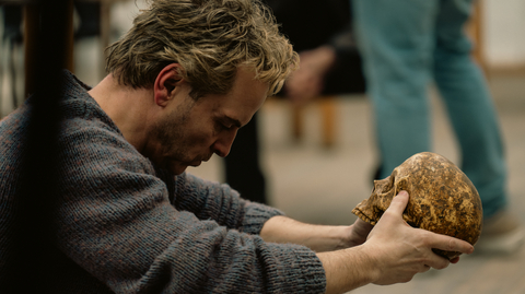 A man sits with a bowed head holding a skull in his hands