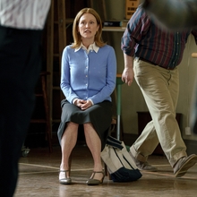 A woman wearing a pale blue cardigan sits on a chair looking straight forward while someone just going out of shot is walking away.