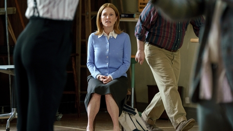 A woman wearing a pale blue cardigan sits on a chair looking straight forward while someone just going out of shot is walking away.