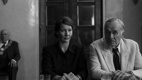A woman and an older man sit next to each other looking forward - the photo is in black and white