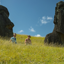 Two men, sitting in a grass field with large rocks behind them