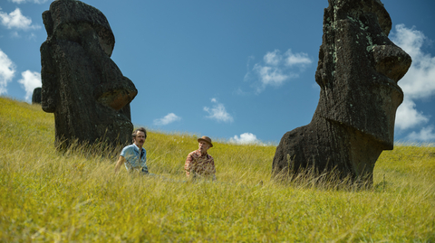 Two men, sitting in a grass field with large rocks behind them