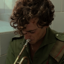 A curly haired person plays a trumpet in what looks like a bathroom