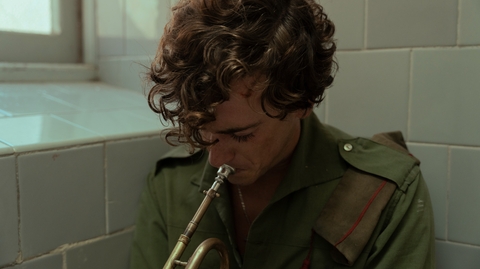 A curly haired person plays a trumpet in what looks like a bathroom