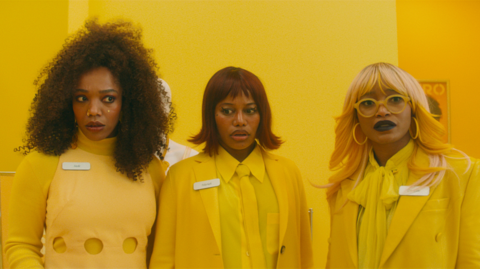 Three people standing in a row looking slightly confused all dressed in yellow against a yellow backdrop
