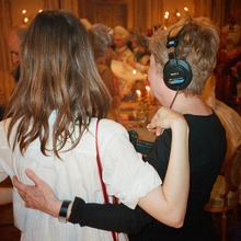 Two people standing with their arms around one another, one of them wearing headphones