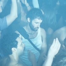 A crowded club dance floor with one person in a white vest looking down with their eyes closed