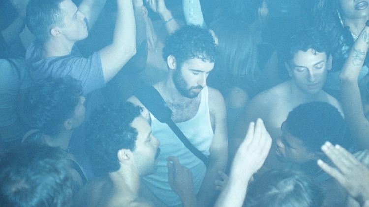 A crowded club dance floor with one person in a white vest looking down with their eyes closed