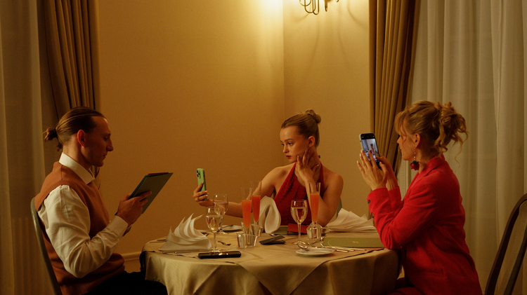 A still of three people sitting at a dinner table all on their phones.