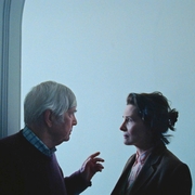 Two people in profile face each other in a dimly lit room with a plain light blue wall in the background.